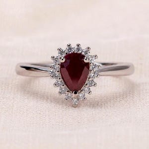 Op de afbeelding: Een zilverkleurige ring met een peervormige, dieprode edelsteen in het midden, omringd door een halo van kleine, heldere, rondgeslepen steentjes. De ringband is glad en gepolijst, wat zorgt voor een klassieke uitstraling met een centrale steen.