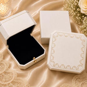 May include: Three white jewellery boxes with a textured finish. One box is open, revealing a black velvet interior and a small gold-toned metal latch. The other two boxes are closed, with one featuring a delicate gold floral border pattern.