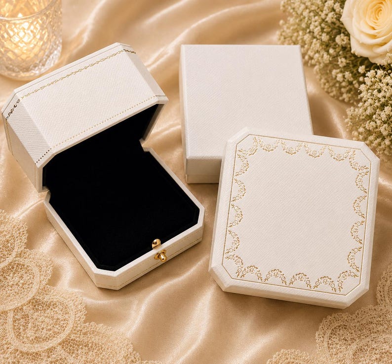 May include: Three white jewellery boxes with a textured finish. One box is open, revealing a black velvet interior and a small gold-toned metal latch. The other two boxes are closed, with one featuring a delicate gold floral border pattern.