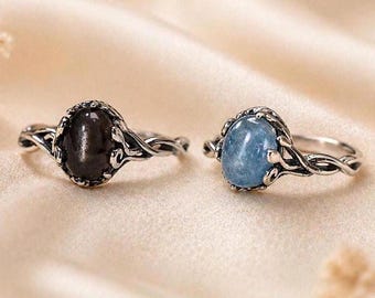 Könnte beinhalten: Zwei Ringe mit komplexen, gedrehten Metallbändern. Der linke Ring hat einen glatten, ovalen schwarzen Stein, während der rechte Ring einen glatten, ovalen hellblauen Stein trägt. Beide Fassungen haben ein dunkles, oxidiertes Finish.