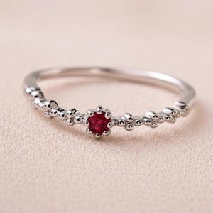 Könnte beinhalten: Ein dünner silberfarbener Ring mit einem kleinen, runden, dunkelroten Edelstein in der Mitte. Das Band ist mit einer Reihe winziger, erhabener Metallkugeln oder Perlen verziert, die sich entlang der Seiten der zentralen Steinfassung erstrecken.