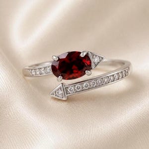 Può includere: Un anello bypass color argento con una gemma centrale di colore rosso intenso dal taglio ovale. La fascia termina con punte a forma di freccia decorate con piccole pietre chiare, e il gambo è impreziosito da ulteriori pietre chiare incastonate.