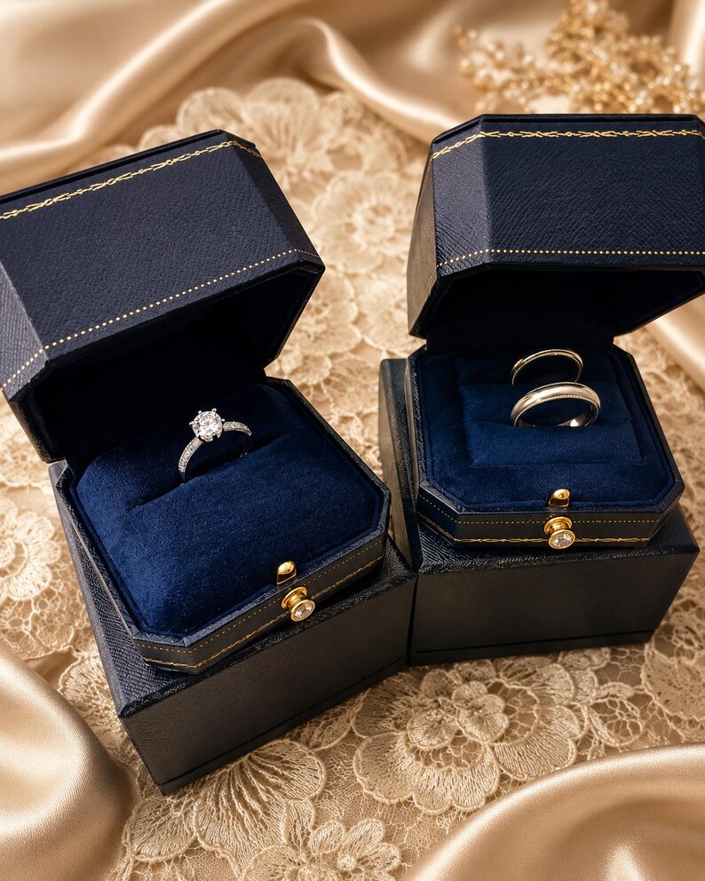 May include: Two dark blue, octagonal jewellery boxes with gold-tone trim and blue velvet interiors. One box holds a silver-toned ring with a central round stone and pav&eacute; accents, while the other box contains two plain silver-toned bands.