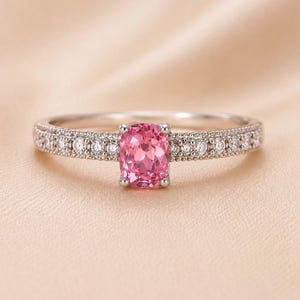 Könnte beinhalten: Ein silberfarbener Ring mit einem zentralen, kissenförmig geschliffenen rosa Edelstein, der von vier Krappen gehalten wird. Die Ringschiene ist auf beiden Seiten mit einer Reihe kleiner, runder, klarer Steine in einer Milgrain-Fassung besetzt.