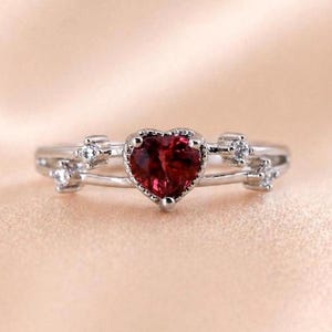 Könnte beinhalten: Ein silberfarbener Ring mit einem zentralen herzförmigen roten Edelstein. Die Ringschiene teilt sich auf beiden Seiten in zwei zarte Arme, die jeweils mit zwei kleinen, klaren, rund geschliffenen Steinen in Metallfassungen besetzt sind.