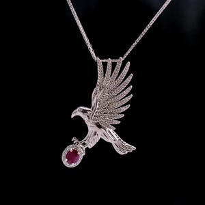 May include: A silver-toned pendant necklace featuring a detailed eagle in flight with its wings spread upwards. The eagle is clutching an oval-shaped, deep red gemstone surrounded by a halo of small, clear stones on a thin silver-toned chain.