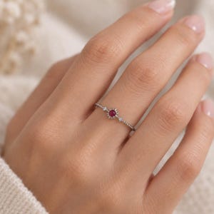 K&ouml;nnte beinhalten: Ein zarter silberfarbener Ring mit einem zentralen runden roten Edelstein in einer sonnenstrahlenf&ouml;rmigen Fassung, flankiert von zwei kleinen klaren Steinen auf einem d&uuml;nnen, perlenartigen Band. Das Design ist symmetrisch gestaltet.
