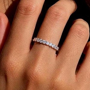 Könnte beinhalten: Ein dünner Metallring mit einer durchgehenden Reihe kleiner, klarer, rund geschliffener Steine auf der oberen Hälfte. Der Ring hat eine polierte Oberfläche und die Steine zeigen ein helles, reflektierendes Funkeln im Licht.