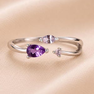 Könnte beinhalten: Ein silberfarbener offener Ring mit drei verschiedenen Edelsteinen. Ein Ende zeigt einen birnenförmigen, tiefvioletten Amethyst, während das andere Ende einen hellrosa Stein im Marquise-Schliff und einen kleinen runden hellrosa Stein trägt.