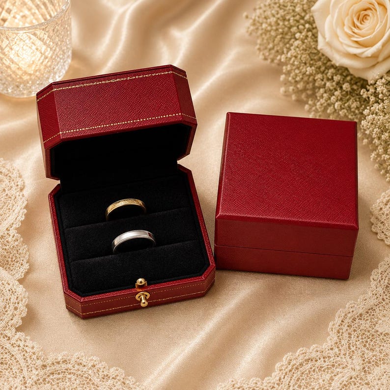 May include: Two dark red, textured jewellery boxes with gold-tone stitching and a gold-tone latch. One box is open, revealing a black velvet interior with two slots holding a gold band and a silver band. The other box is closed and sits beside it.