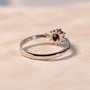 K&ouml;nnte beinhalten: Ein silberfarbener Ring mit einer kreisf&ouml;rmigen Fassung und einem tiefroten Edelstein in der Mitte. Die Fassung ist von einer sonnenstrahlenartigen Anordnung kleiner, klarer, facettierter Steine umgeben. Die Ringschiene ist glatt und poliert mit einer &Ouml;ffnung.