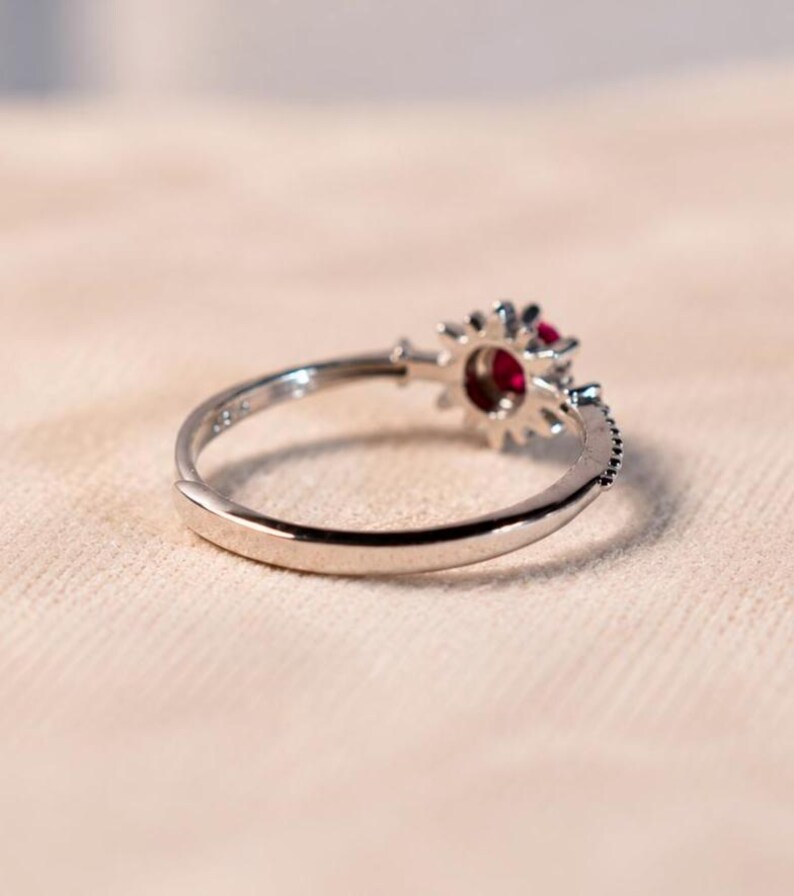 K&ouml;nnte beinhalten: Ein silberfarbener Ring mit einer kreisf&ouml;rmigen Fassung und einem tiefroten Edelstein in der Mitte. Die Fassung ist von einer sonnenstrahlenartigen Anordnung kleiner, klarer, facettierter Steine umgeben. Die Ringschiene ist glatt und poliert mit einer &Ouml;ffnung.