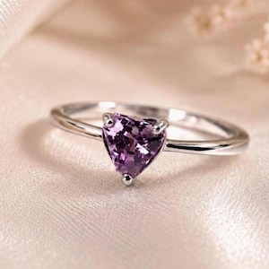Könnte beinhalten: Ein silberfarbener Ring mit einem einzelnen herzförmigen violetten Edelstein in einer Krappenfassung. Die Ringschiene ist dünn und poliert und weist eine glatte metallische Oberfläche auf, die den facettierten violetten Stein ergänzt.