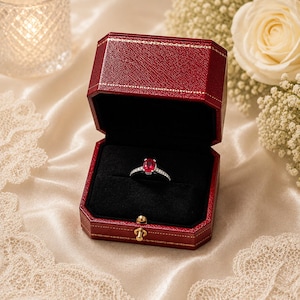 May include: A silver ring featuring an oval-cut red gemstone set in a dark red, octagonal, hinged jewellery box with gold-tone trim and a black velvet interior lining, presented in an open position to display the ring clearly.
