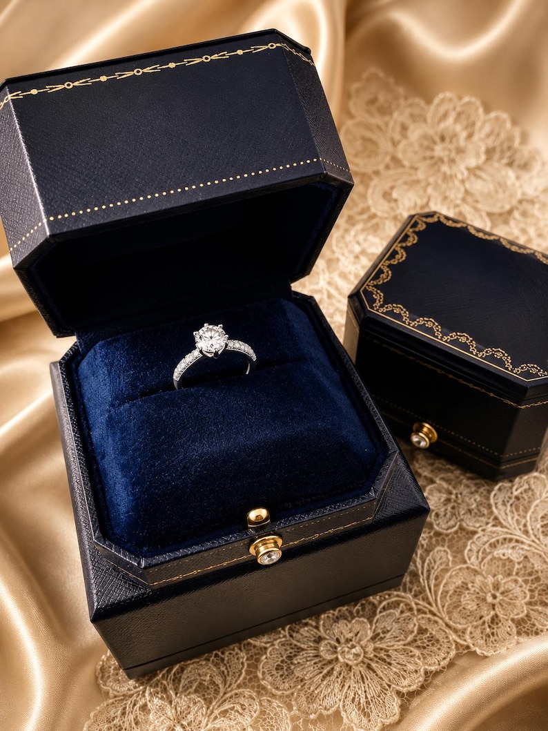 May include: A silver-coloured ring with a central round-cut stone and smaller stones along the band, presented inside an open, dark blue, hexagonal jewellery box with gold-coloured trim and a small circular clasp, alongside a closed matching box.