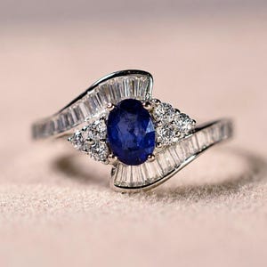 Könnte beinhalten: Ein silberfarbener Ring mit einem zentralen, oval geschliffenen blauen Saphir. Die Ringschiene ist mit Reihen von klaren Steinen im Baguetteschliff sowie zwei Clustern aus je drei klaren Steinen im Rundschliff an den Seiten verziert.