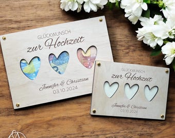 May include: Two wooden boxes with heart-shaped cutouts. The top box has three hearts with money visible through the cutouts. The bottom box has three empty hearts. Both boxes have the text "Glückwunsch zur Hochzeit" and the names "Jennifer & Christian" with the date "03.10.2024".