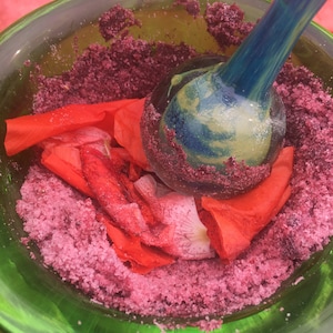 Puede incluir: Un bol de vidrio verde lleno de cristales de azúcar rosa y pétalos de flores rojas. Un mortero de vidrio azul descansa en el bol.