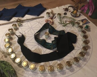 May include: A white cloth with a flower of life pattern, surrounded by small glass jars with gold lids. There are two black and blue sashes, a wooden stick, and a small antler on the cloth.
