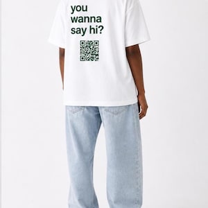 Könnte beinhalten: Weißes T-Shirt mit dem grünen Text "you wanna say hi?" und einem QR-Code. Das Model trägt hellblaue Jeans. Das T-Shirt ist ein lässiges Kleidungsstück für den Alltag.