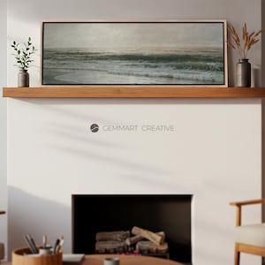 May include: A framed seascape painting with a light blue and green color scheme depicting a stormy ocean with waves crashing on the shore. The painting is hanging on a wooden shelf above a fireplace.