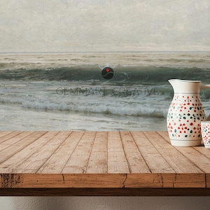 May include: A wooden table with a white pitcher and a white mug with red and green floral patterns. The pitcher and mug are on a wooden table in front of a painting of a seascape.