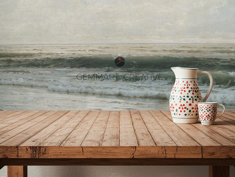 May include: A wooden table with a white pitcher and a white mug with red and green floral patterns. The pitcher and mug are on a wooden table in front of a painting of a seascape.