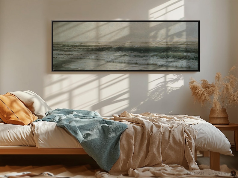 May include: A bedroom with a bed made with a light brown blanket and a light blue throw blanket. A black framed picture of a seascape hangs on the wall above the bed. A vase with dried pampas grass sits on a table in the corner of the room.