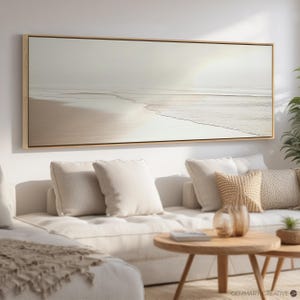 Pode incluir: Uma impressão em tela horizontal e longa de uma cena de praia com areia branca, água azul e um céu azul claro. A impressão é emoldurada em uma moldura de madeira clara.