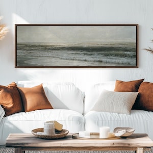 May include: A long, horizontal framed print of a seascape with waves crashing on the shore. The print is hanging on a white wall above a white couch with brown throw pillows.