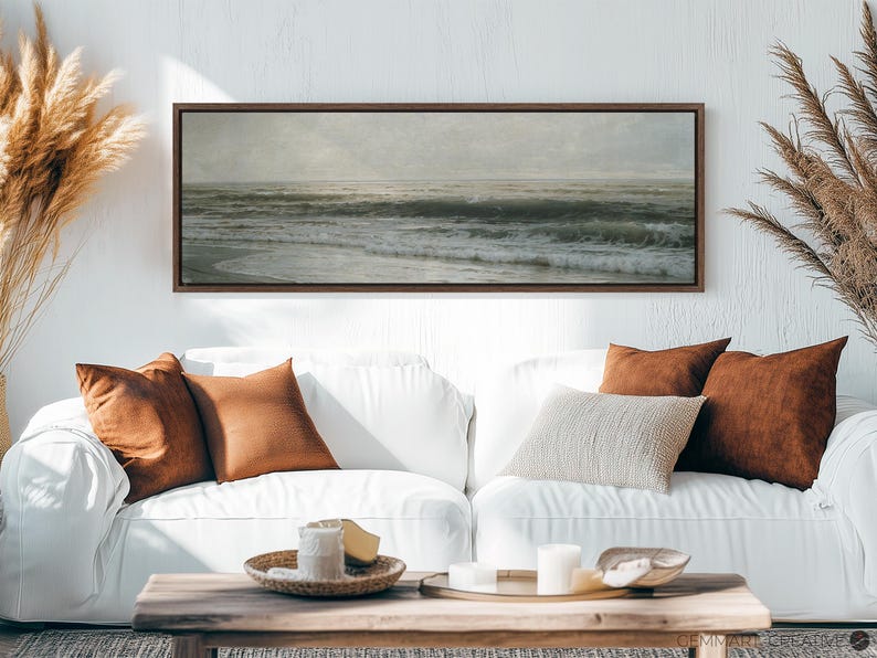 May include: A long, horizontal framed print of a seascape with waves crashing on the shore. The print is hanging on a white wall above a white couch with brown throw pillows.
