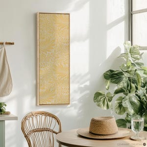 May include: A framed print with a yellow floral pattern hangs on a white wall. The print is in a wooden frame and has a light yellow background with a darker yellow floral design. The print is hanging in a room with a wicker chair and a potted plant.
