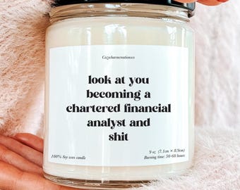 May include: A glass jar candle with a black lid, held by a person. The candle has a white label with the text "look at you becoming a chartered financial analyst and shit." It is 9 oz (7.1cm x 8.9cm) and burns for 50-60 hours.