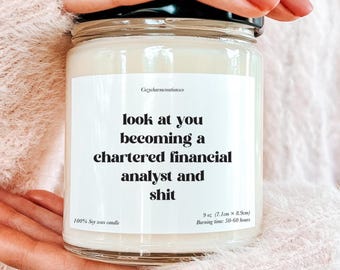 May include: A clear glass jar candle with a black lid, held by a person. The candle has a white label with the text "look at you becoming a chartered financial analyst and shit." The candle is 9 oz and 7.1cm x 8.9cm.