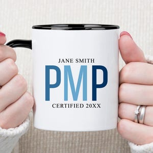 May include: White ceramic mug with a black rim and handle, featuring the name "Jane Smith" and the letters "PMP" in blue, with "CERTIFIED 20XX" below. The mug is held by hands wearing rings.