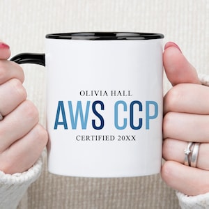May include: White ceramic mug with a black rim and handle, featuring the text "OLIVIA HALL AWS CCP CERTIFIED 20XX" in blue and black lettering. The mug is held by a person wearing a white jumper.