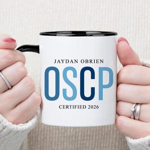 Puede incluir: Taza de cerámica blanca con borde y asa negros, con el nombre "JAYDAN OBRIEN" sobre el texto azul "OSCP" y el texto "CERTIFIED 2026". La taza es sostenida por una persona que lleva un suéter blanco.