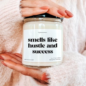 May include: A white soy candle with a black label that reads "smells like hustle and success". The candle is 9 oz and has a burn time of 50-60 hours.
