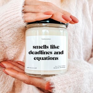Puede incluir: Una vela de soja blanca con una etiqueta negra que dice "smells like deadlines and equations". La vela pesa 9 oz y tiene un tiempo de combustión de 50-60 horas.