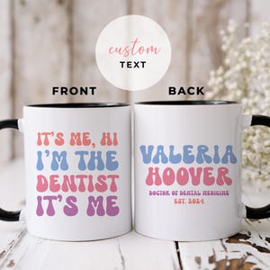 Puede incluir: Taza de café de cerámica blanca con borde y asa negros. La parte delantera de la taza tiene el texto "It's me, hi I'm the dentist It's me" en rosa, azul y azul claro. La parte posterior de la taza tiene el texto "Valeria Hoover Doctor of Dental Medicine Est. 2024" en azul, rosa y azul claro.