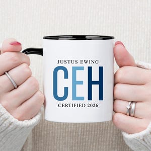May include: White ceramic mug with a black rim and handle, featuring the text "JUSTUS EWING CEH CERTIFIED 2026" in blue and black. The mug is held by a person wearing a white sweater.