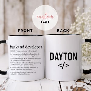 Op de afbeelding: Een witte keramische koffiemok met een zwarte rand en handvat. De voorkant van de mok heeft de definitie van "backend developer" in zwarte tekst gedrukt. De achterkant van de mok heeft de naam "DAYTON" in zwarte tekst met haakjes gedrukt.