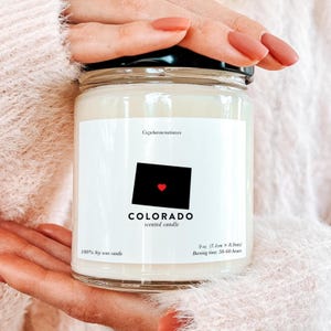 Op de afbeelding: Een heldere glazen potkaars met een zwart deksel. Het etiket toont een zwarte omtrek van de staat Colorado met een rood hart, het woord "COLORADO" en "scented candle". De kaars is 7,1 cm x 8,9 cm en brandt 50-60 uur.