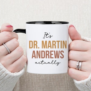 May include: White ceramic mug with a black rim and handle. The mug has the text "It's Dr. Martin Andrews actually" printed on it in black and gold lettering.