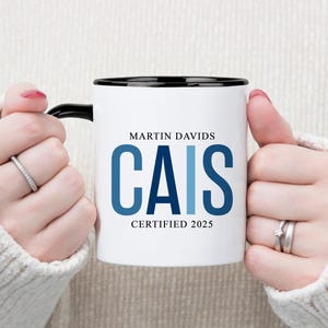 May include: White ceramic mug with a black rim and handle. The mug features the name "MARTIN DAVIDS" above the word "CAIS" in blue, with "CERTIFIED 2025" below. The mug is held by a person wearing a white sweater.
