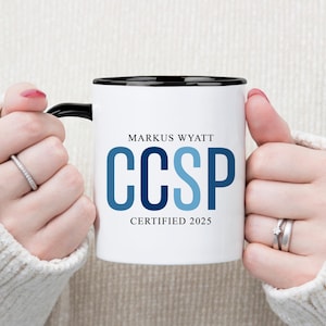 May include: White ceramic mug with a black rim and handle, featuring the text "Markus Wyatt CCSP CERTIFIED 2025" in blue. The mug is held by hands wearing rings, against a neutral background.