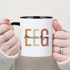 Op de afbeelding: Witte keramische mok met een zwarte rand en handvat. De mok heeft de letters "EEG" in een groot, kleurrijk lettertype met de naam "Maritza" eronder in cursief.