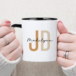 May include: White ceramic mug with a black rim and handle, featuring the initials "JD" in a warm brown and beige color scheme, with the name "Madilynn" in a handwritten font. The mug is held by a person.