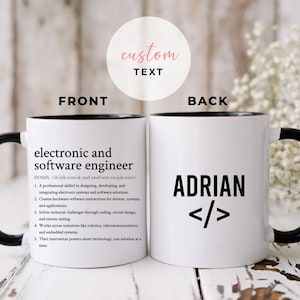 May include: White ceramic mug with a black handle and rim. The front of the mug features the text "electronic and software engineer" with a definition. The back of the mug displays the name "ADRIAN" and code symbols.