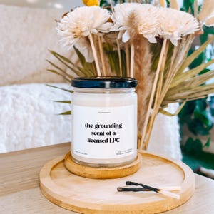 a candle labeled "the grounding scent of a licensed LPC" placed on a wooden tray, with a vase of white flowers in the background.
